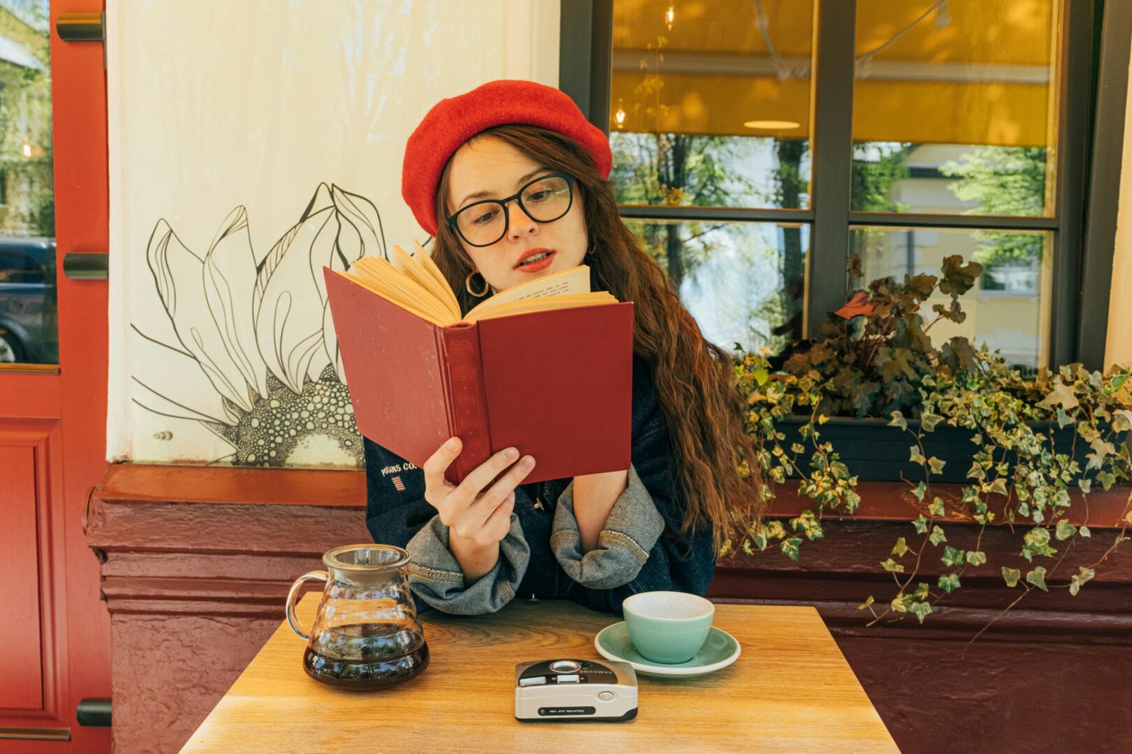 El propósito de la lectura en 2026: "De Lector a Creador en una tarde" 4 "Mujer con boina roja y gafas sentada en café leyendo un libro rojo abierto; representa la concentración y el propósito de lectura."